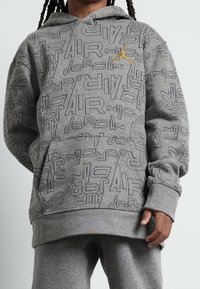 Graue Kapuzenjacke mit einem durchgehenden grafischen Muster, das die Worte "AIR" und "JORDAN" zeigt. Enthält eine Kängurutasche und eine Ziehkordel an der Kapuze.