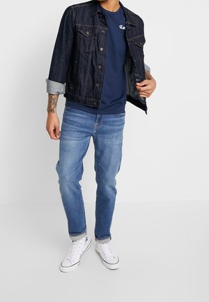 Veste en denim bleu foncé, superposée sur un t-shirt marine, associée à un jean bleu clair et des baskets montantes blanches.