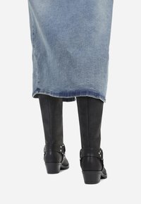 Botas negras hasta la rodilla con una textura suave, que presentan un detalle de correa y hebilla en el tobillo, combinadas con una falda de mezclilla.