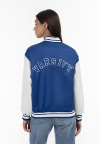 Giacca bomber varsity blu con maniche bianche, con la scritta "VARSITY" sulla schiena. Ha un colletto e polsini a coste con righe blu e bianche.