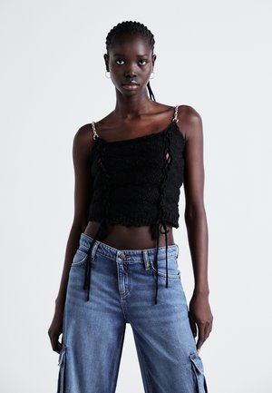 Melna adīta cropped tops ar regulējamiem saitēm un sudraba ķēdes siksnām, apvienots ar gaiši ziliem platām kājām džinsiem ar kabatām un pogu aizdari.