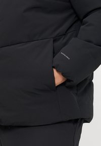Black puffer jacket with a textured finish, featuring a side pocket and branding on the sleeve. The fabric is smooth and matte.