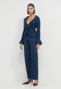Femme portant un pull en maille côtelée bleu marine à col en V profond avec des manches évasées, un jean bleu à jambes larges orné de perles blanches, et des chaussures à talons noires.