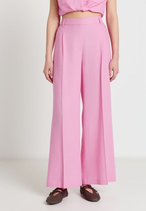 Femme portant un pantalon rose taille haute à jambes larges et des chaussures plates tressées marron, debout devant un fond clair uni.