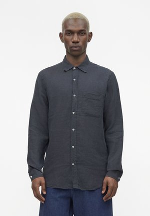 Muž s krátkymi blond vlasmi, oblečený v tmavú košeľu na gombíky s náprsným vreckom a modré džínsy, stojaci pred jednoduchým pozadím.