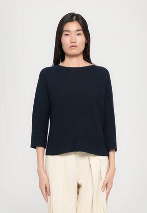 Navy blue, ribbed knit sweater with a rounded neckline and three-quarter sleeves. Hem is slightly curved. Paired with light-colored pleated trousers.