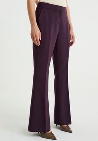 Pantaloni a zampa d'elefante in tessuto viola scuro, con una texture liscia e una vita aderente. Indossati con scarpe a punta.