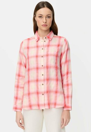 Kobieta ubrana w koszulę na guziki w różowo-białą kratę z długim rękawem i kieszenią na piersi, w połączeniu z jasnymi spodniami, na jednolitym tle.