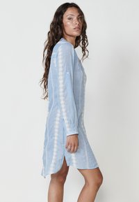 Helles blaues Kleid mit langen Ärmeln, das weiße dekorative Stickerei entlang der Seiten und einen geschwungenen Saum aufweist. Hergestellt aus einem leichten Stoff.