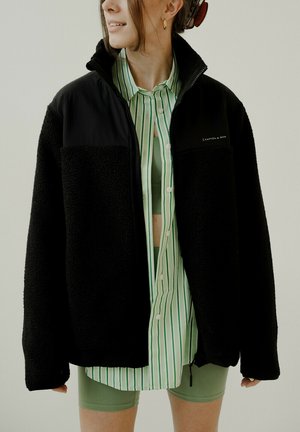 Giacca in pile nera con materiale testurizzato, chiusura con zip e collare alto, indossata sopra una camicia a righe verde e bianca e pantaloni corti verde chiaro.