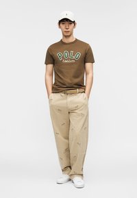 Καφέ κοντομάνικο μπλουζάκι με το λογότυπο "POLO", μπεζ παντελόνι με κεντημένες λεπτομέρειες, λευκά αθλητικά παπούτσια και λευκό καπέλο.