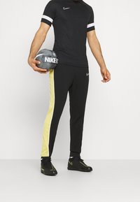 Schwarzes Nike-T-Shirt mit weißen Ärmelstreifen, kombiniert mit schwarzen Hosen, die einen gelben Seitenstreifen haben. Hält einen grauen Fußball.