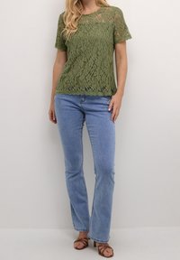 Haut en dentelle vert olive à manches courtes, ourlet festonné et motif floral, associé à un jean droit bleu clair et des sandales marron.