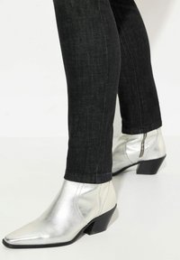 Botines metálicos plateados con puntera afilada y tacón bajo, combinados con jeans skinny oscuros. Los botines presentan un detalle de cremallera lateral.