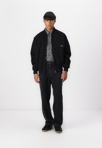 Black bomber jacket with striped shirt underneath, dark jeans, and black sneakers. Features ribbed cuffs and zip closure on the jacket.