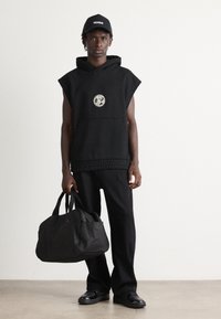 Man wearing black sleeveless hoodie, loose black pants, black cap, and shoes, holding a large black duffel bag, standing on a plain background.