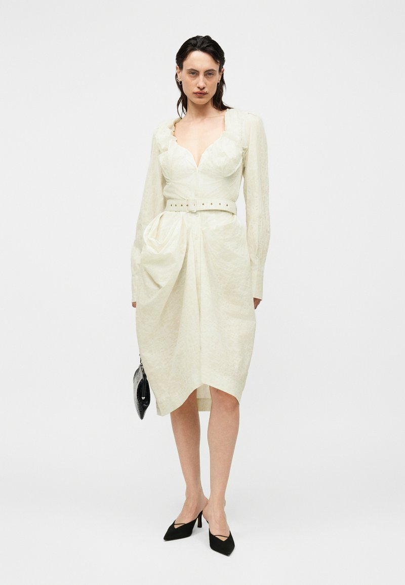 Femme portant une robe écrue ceinturée à manches longues, tenant un sac à main noir et portant des escarpins noirs à bouts pointus.