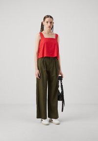 Červený lněný crop top se čtvercovým výstřihem, doplněný olivově zelenými širokými kalhotami. Model drží černou kabelku; vzhled doplňují bílé tenisky.