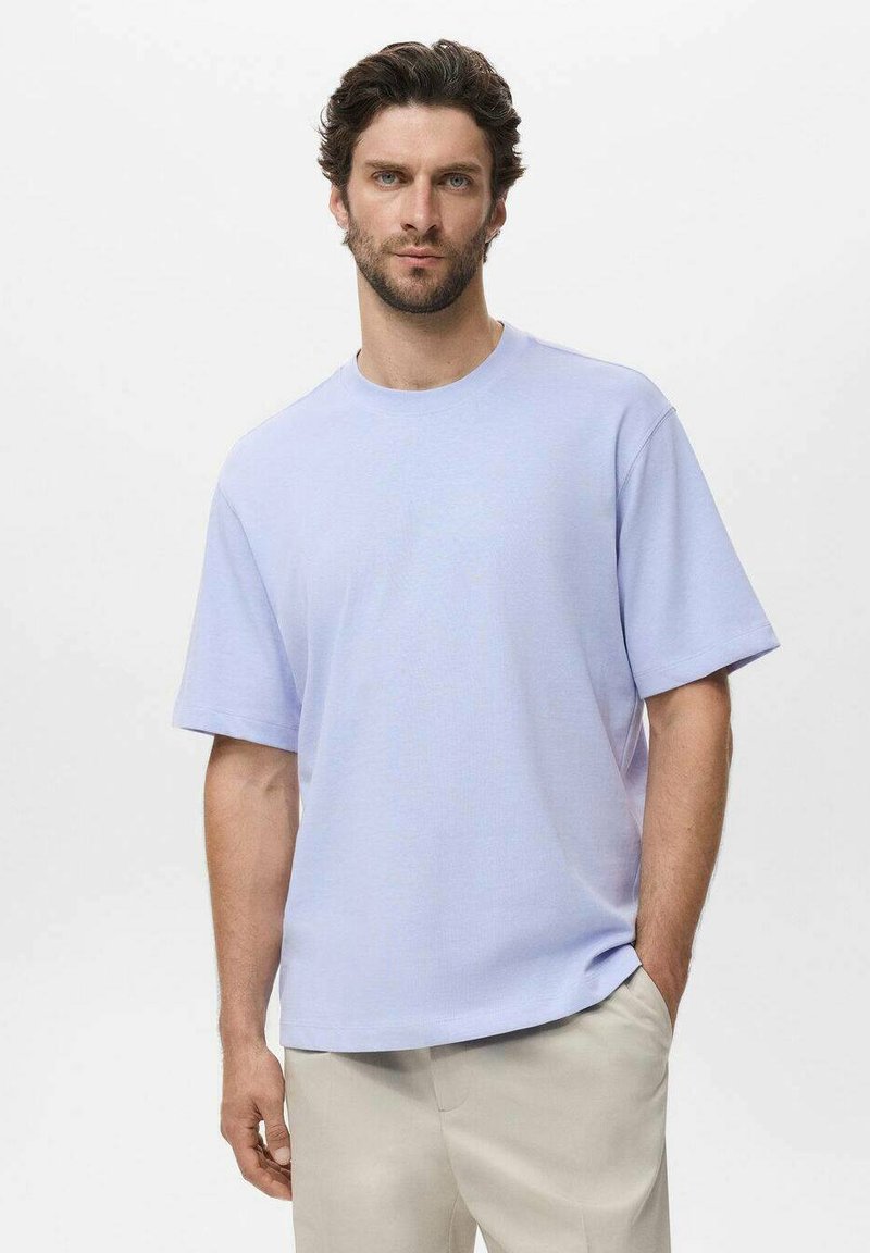 Tílko s krátkým rukávem v levandulové barvě, vyrobené z měkké látky, s volným střihem a kulatým výstřihem. Nosené s světlými kalhotami.