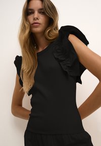 Černý žebrovaný top s texturovanými volánovými rukávy, slim fit střihem a kulatým výstřihem, který zvýrazňuje detaily v materiálu a celkovém designu.