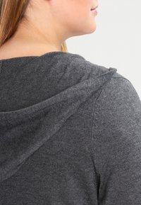 Szary, teksturowany sweter z kapturem z widocznym szwem biegnącym wzdłuż pleców. Kaptur jest miękki i naturalnie opada.