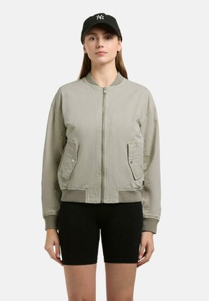 Femme portant une veste bomber beige zippée, un cuissard de cyclisme noir et une casquette de baseball noire avec un logo blanc, debout devant un fond uni.
