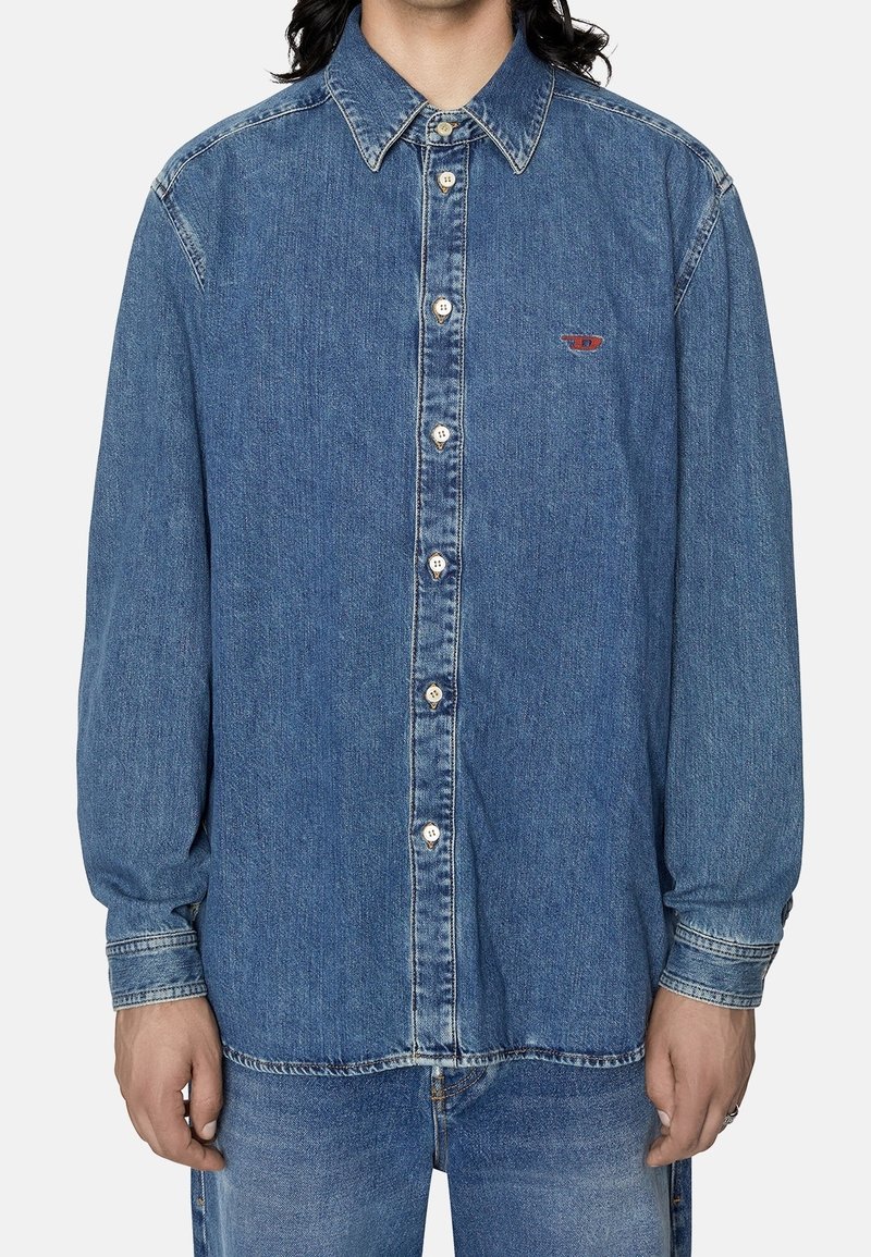 Camicia in denim di un blu medio con maniche lunghe, colletto a bottoni e placca frontale con bottoni. Presenta un piccolo logo rosso sopra la tasca sinistra.