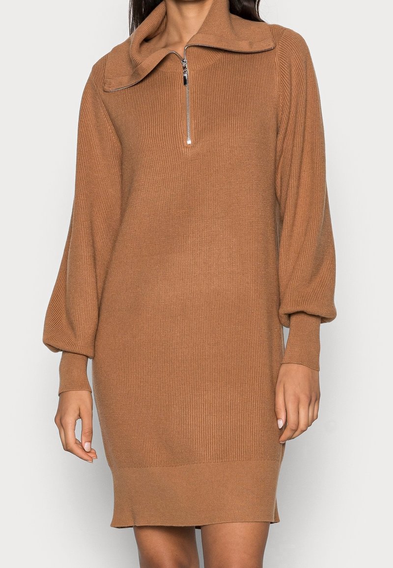 Femme portant une robe tricotée côtelée couleur camel avec un zip à quart et des manches ballon, se tenant contre un fond blanc uni.