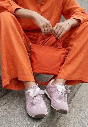Persoon in een oranje outfit zittend op stenen trappen met een kleine oranje handtas, draagt lichtpaarse sneakers met witte veters en het merk "Venezia".