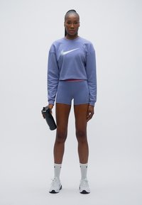 Μωβ cropped φούτερ με λογότυπο Nike, σε συνδυασμό με ανοιχτό μωβ σορτς. Το μοντέλο κρατάει ένα μαύρο παγούρι νερού, φοράει λευκές κάλτσες και αθλητικά παπούτσια.