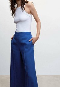 Kobieta w bezrękawnikowej białej bluzce i wysokich, szerokich niebieskich spodniach w kolorze królewskim, z rękami w kieszeniach na jednolitym tle.