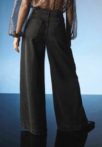 Mustat leveälahkeiset farkut korkealla vyötäröllä, joissa on kaksi takataskua ja sileä pinta, yhdistettynä läpikuultavaan, paljettiyksityöllä varustettuun toppiin.