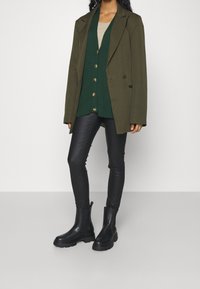 Olivgrön oversized kavaj över en mörkgrön knappad cardigan, ihop med svarta skinnybyxor och svarta ankle boots. Släta texturer genomgående.