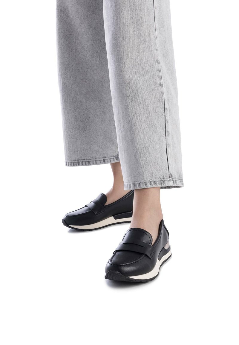 Zapatos negros de cuero tipo mocasín con punta redondeada, detalle de tira de cuero, suela blanca acolchada y suela texturizada. Se llevan con unos vaqueros de pierna ancha de color gris claro.