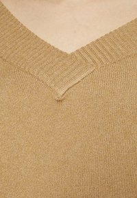 Pull à col en V dans un ton beige chaud, avec des bords côtelés et un tricot doux et texturé. Gros plan sur l'encolure et les détails du tissu.