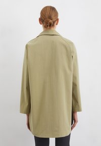 Palton lung de culoare verde măsliniu, cu guler, mâneci trei sferturi și croială dreaptă, având o textură netedă și slituri laterale.