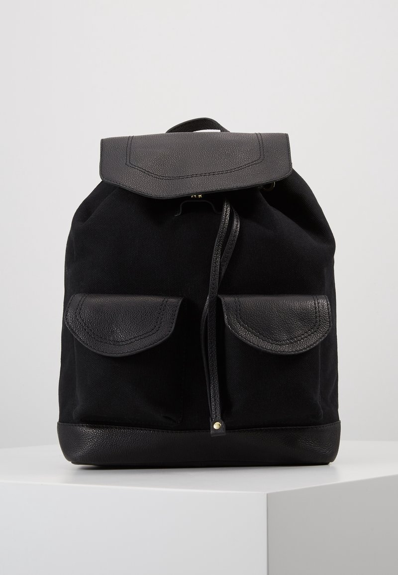 Sac à dos en tissu noir avec rabat en cuir, deux poches avant avec rabats en cuir, fermeture à cordon et fond en cuir sur une surface blanche.