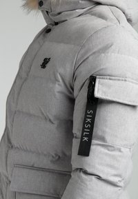 Graue Pufferjacke mit gestepptem Design, ausgestattet mit einer fellgefütterten Kapuze, Logo auf der Brust und einer Reißverschlusstasche am Arm.