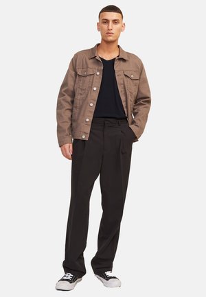 Junger Mann steht mit einer Hand in der Tasche, trägt eine braune, geknöpfte Jacke, ein schwarzes Hemd, schwarze Hose und schwarz-weiße Turnschuhe.