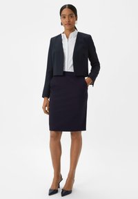 Marineblauwe cropped blazer met lange mouwen over een witte coltrui, gecombineerd met een bijpassende marineblauwe kokerrok en hakken met een spitse neus.