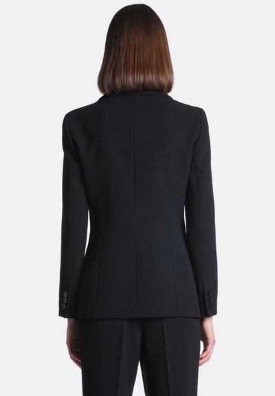 Femme aux cheveux bruns lisses mi-longs, portant un blazer noir ajusté et un pantalon noir assorti, vue de dos sur fond blanc.