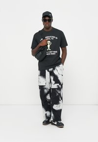 Svart grafisk t-shirt med vit text och utomjording-design, kombinerad med svart-vita mönstrade cargobyxor. Accessoarer inkluderar en keps och solglasögon.