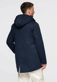 Parka à capuche bleu marine avec une coupe droite, dotée d'un ourlet fendu et d'une texture lisse. Design simple avec des accents matériels minimalistes.
