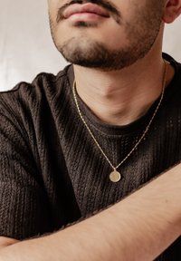 Ciondolo circolare in oro su una delicata catena indossato sopra un maglione scuro e texturizzato. Il ciondolo presenta una superficie liscia e un bordo sottile.