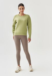 Suéter verde claro con la palabra "BALANCE" en relieve, combinado con leggings beige y zapatillas neutras. Textura suave, corte relajado y diseño minimalista.