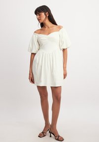 Biała sukienka odsłaniająca ramiona, z bufkowymi rękawami, marszczoną górą i rozkloszowanym dołem. Gładki materiał z delikatnym połyskiem, bez wzorów i ozdób.