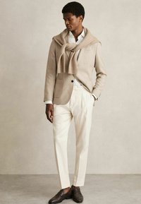 Sacou bej cu buzunar, cămașă albă, pantaloni crem și pantofi loafer maro. Pulover aruncat pe umeri, creând un look stratificat. Texturi netede.