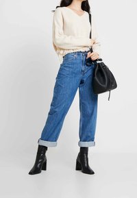 Kremowy sweter z dekoltem w serek i falbaniastym dołem, połączony z podwiniętymi niebieskimi dżinsami oraz czarnymi botkami do kostek. W ręku trzymana czarna torba typu bucket.