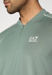 Πράσινο αθλητικό τοπ με λαιμόκοψη μισού φερμουάρ, υφή από δίχτυ και λευκό λογότυπο. Διαθέτει διακριτική λευκή λεπτομέρεια σε καθαρό χρώμα κατά μήκος των ώμων.