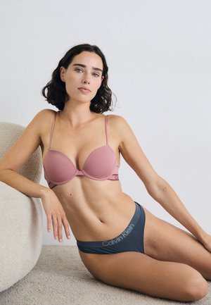 Une femme aux cheveux courts et ondulés est assise sur un canapé rond et moelleux, portant un soutien-gorge rose et un sous-vêtement gris Calvin Klein, regardant la caméra avec confiance.